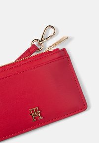 Piros bőrtárca texturált felülettel, aranyszínű cipzárral és kiegészítőkkel, valamint arany színű logós részlettel az elején.