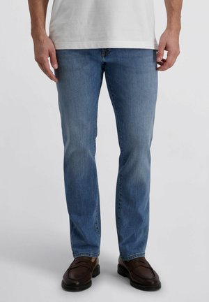 Mann trägt hellblaue Jeans, braune Lederschuhe und ein weißes Hemd, steht vor einem einfarbigen hellen Hintergrund.