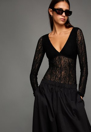 Fekete csipkés body hosszú ujjakkal és mély V-nyakkivágással, fekete magas derekú szoknyával párosítva. Sima anyag; áttetsző részletekkel.