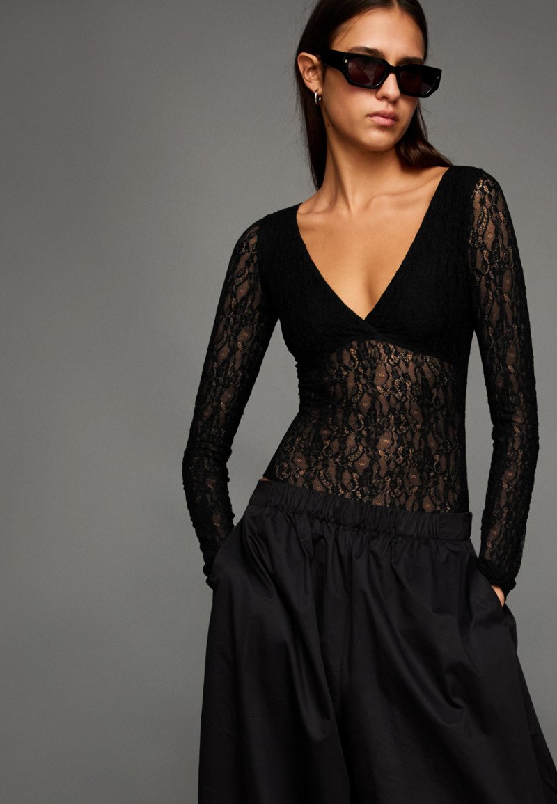 Čierny čipkovaný body s dlhými rukávmi a hlbokým výstrihom do V, skombinovaný s čiernou sukňou s vysokým pásom. Hladká látka; priehľadné detaily.