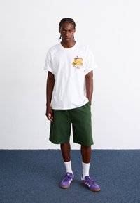 White short-sleeve t-shirt with a printed leopard design, paired with green shorts, white socks, and purple sneakers on a blue carpet background.