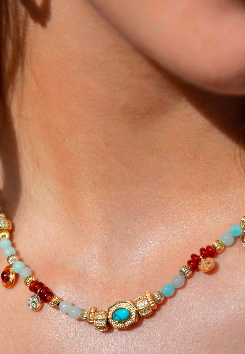 Collier en perles avec des accents dorés, comportant des perles turquoises, rouges et vertes, et des détails complexes. Surface lisse, vue rapprochée sur la peau.