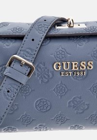 Mėlyna dirbtinės odos rankinė su reljefiniais raštais, sidabrinėmis detalėmis ir auksiniu "GUESS EST 1981" logotipu. Priklausomas diržas yra komplekte.