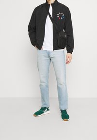 Svart lättviktsjacka med flerfärgade broderade löv, vit t-shirt, ljusblå jeans och gröna sneakers med vita ränder.