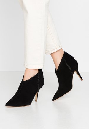 Femme portant des bottines en daim noir à bout pointu avec talons aiguilles et un pantalon blanc raccourci, sur un fond uni.
