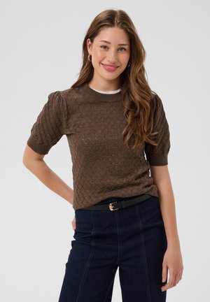 Jeune femme aux longs cheveux ondulés portant un pull marron texturé à manches courtes, pantalon bleu foncé avec ceinture noire, souriante avec la main sur la hanche.