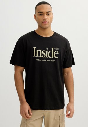 Joven vestido con una camiseta negra con el texto "Inside Where Passion Meets Work" y pantalones cargo beige, de pie sobre un fondo blanco liso.