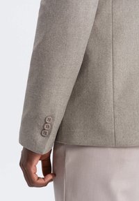 Blazer ajusté gris clair en tissu texturé, avec un tissage subtil et trois boutons marron sur le poignet, présenté de côté.