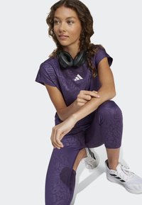 Young woman in purple athletic wear and white sneakers kneeling, with black headphones resting around her neck, looking to the side.