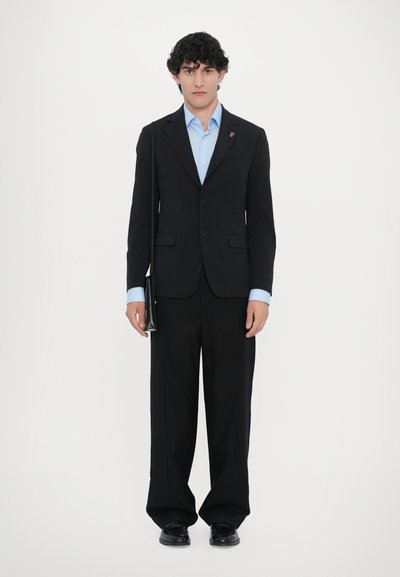 Schwarzer maßgeschneiderter Anzug mit einem blauen Hemd und einem Einstecktuch. Weit geschnittene Hose, glattes Material und schwarze formelle Schuhe. Hält eine schwarze Tasche.
