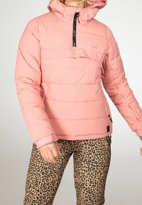 Personne portant une veste matelassée rose avec une fermeture éclair noire et un pantalon à motifs léopard, debout devant un fond uni.