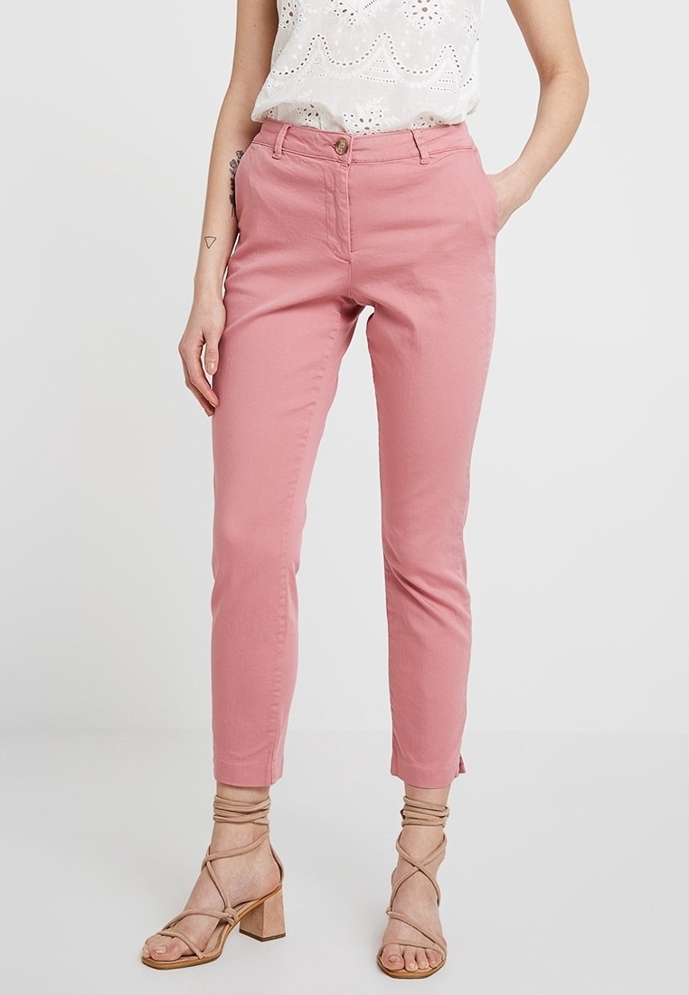 Femme portant un pantalon slim rose taille haute, un top blanc sans manches à œillets, et des sandales beige à talons blocs avec des brides, mains dans les poches.