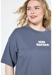 Šed�é oversized tričko s bílým nápisem "ICED MATCHA." Vyrobeno z měkké tkaniny s uvolněným střihem a kulatým výstřihem.