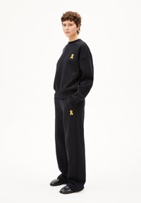Zwarte sweatshirt en broek van zacht materiaal, met een klein geel lintlogo op de borst en de zak. Ontwerp met een losse pasvorm. Zwarte schoenen.