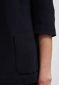 Námořnický modrý kabát s texturovaným povrchem, s nenápadnou kapsou na levé straně a mírně ohrnutým rukávem.