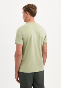 Man die met zijn rug naar de camera staat, draagt een lichtgroen overhemd met korte mouwen en een donkere broek, tegen een effen witte achtergrond.