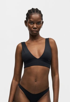 Femeie cu părul împletit purtând un top de bikini negru cu decolteu adânc în V și chiloți de bikini asortați, cu talie joasă, pe un fundal simplu și deschis.