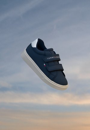 Mørkeblå sneakers med hvid sål og velcrolukninger, Tommy Hilfiger-logo på remmen og siden, svævende mod en skyet himmelbaggrund.