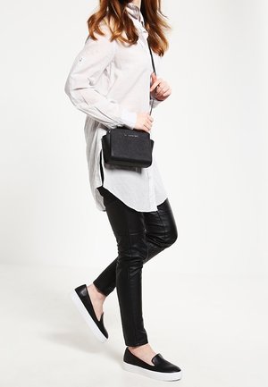 Borsa a tracolla nera con superficie strutturata, accessori in argento, indossata con una camicia lunga bianca con bottoni e pantaloni neri simili alla pelle. Scarpe nere senza lacci.