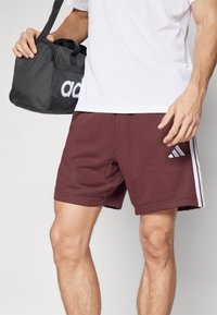 Burgundy idrottsshorts i mjukt material, med vita sidostripes och en Adidas-logotyp. Kombineras med en svart sportväska.