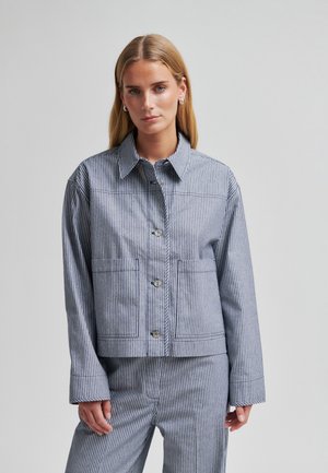 Kvinde iført en grå og hvid lodret striped knap-op jakke og matchende bukser, stående mod en ensfarvet lys grå baggrund.