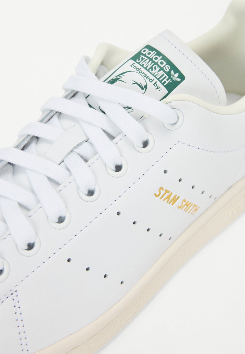 Sneaker en cuir blanc avec des détails perforés, lacets blancs plats, et texte doré "STAN SMITH". Présente un logo vert et une semelle beige.
