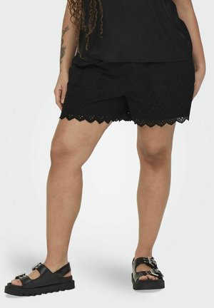 Shorts noirs avec un ourlet festonné et des motifs en œillets, associés à des sandales noires avec des semelles épaisses et des boucles décoratives.