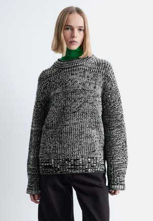 Schwarz-weiß gemusterter Pullover mit legerer Passform, gerippten Bündchen und rundem Ausschnitt, kombiniert mit einem grünen Rollkragenoberteil.