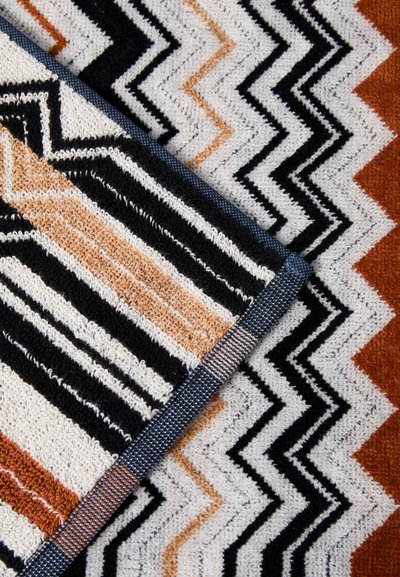Textil szőnyeg, amelyen cikkcakk mintázatok találhatók fekete, fehér, narancs és barna színekben. Puha, textúrált anyag tartós szőtt szélekkel.