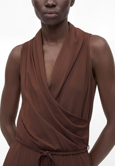 Femme portant une robe portefeuille marron sans manches avec un décolleté en V profond et une fine ceinture nouée à la taille, debout devant un fond blanc uni.