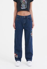 Blue denim wide-leg jeans featuring embroidered designs on each leg. Paired with a black cropped t-shirt. White sneakers complete the look.