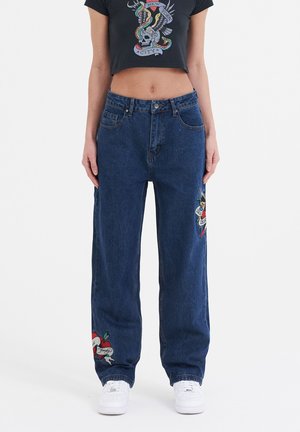 Blaue Denim-Jeans im weit geschnittenen Stil mit gestickten Motiven auf jedem Bein. Kombiniert mit einem schwarzen, kurz geschnittenen T-Shirt. Weiße Sneakers runden den Look ab.