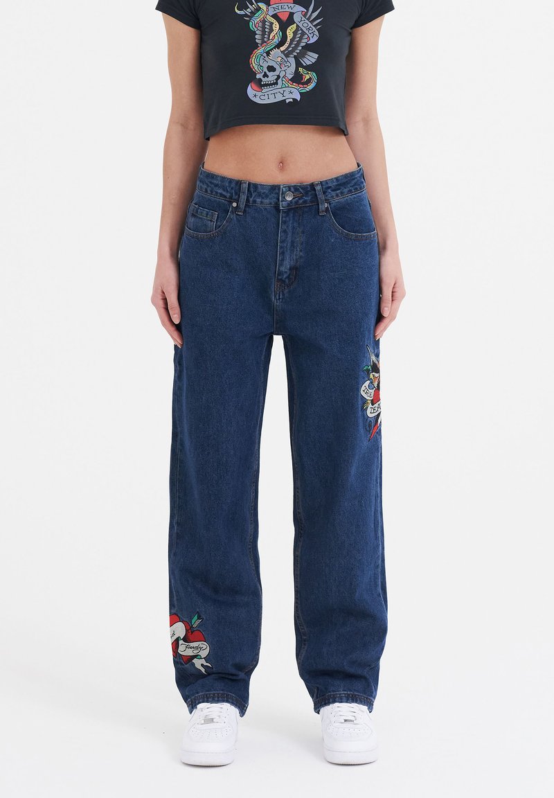 Blue denim wide-leg jeans featuring embroidered designs on each leg. Paired with a black cropped t-shirt. White sneakers complete the look.