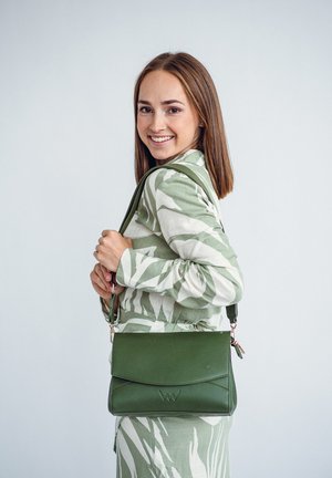 Zelená kožená kabelka s klapkou, zdobená texturovanými akcenty a odnímatelným ramenním popruhem. Nesená s vzorovaným zeleným outfitem.