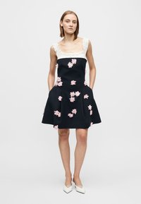 Vestito nero con scollo arricciato bianco e ricami floreali rosa. Linea ad A con tasche, tessuto liscio e lunghezza al ginocchio.