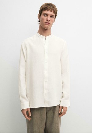 Mladý muž nosící bílou košili s dlouhým rukávem, knoflíky a límečkem typu band a hnědé kalhoty, stojí před jednobarevným světlým pozadím.