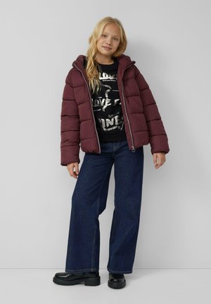 Blondine in einer kastanienbraunen Daunenjacke, schwarz bedrucktem Sweatshirt, weit geschnittenen dunklen Jeans und schwarzen klobigen Stiefeln, steht vor einer schlichten Wand.