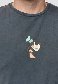 Donkergrijze katoenen t-shirt met daarop een kleurrijk cartoonfiguur van een hond die een koksmuts draagt, gepositioneerd op de linkerkant van de borst.