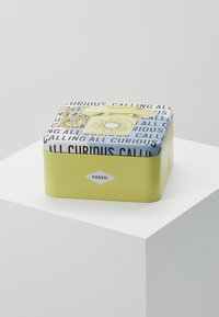 Boîte en fer jaune Fossil avec un téléphone rotatif vintage et le texte « Calling All Curious » sur le couvercle, placée sur un socle blanc.