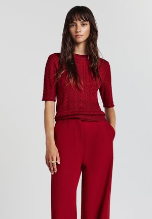 Donna con lunghi capelli castani, che indossa un maglione a maniche corte lavorato a maglia di colore rosso e pantaloni a vita alta rossi abbinati, in piedi con una mano in tasca.