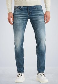 Jeans de denim azul ajustados con un lavado desgastado, sutiles arrugas, diseño clásico de cinco bolsillos y detalles de costura en contraste. Combinados con zapatillas blancas.