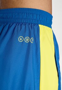 Niebieskie i żółte spodenki sportowe z elastycznym pasem, wykonane z teksturowanej tkaniny, posiadające boczne kieszenie oraz nadrukowane numery na niebieskiej części.