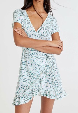 Woman wearing a white wrap dress with blue polka dots and ruffled hem, standing with arms crossed against a plain background.
