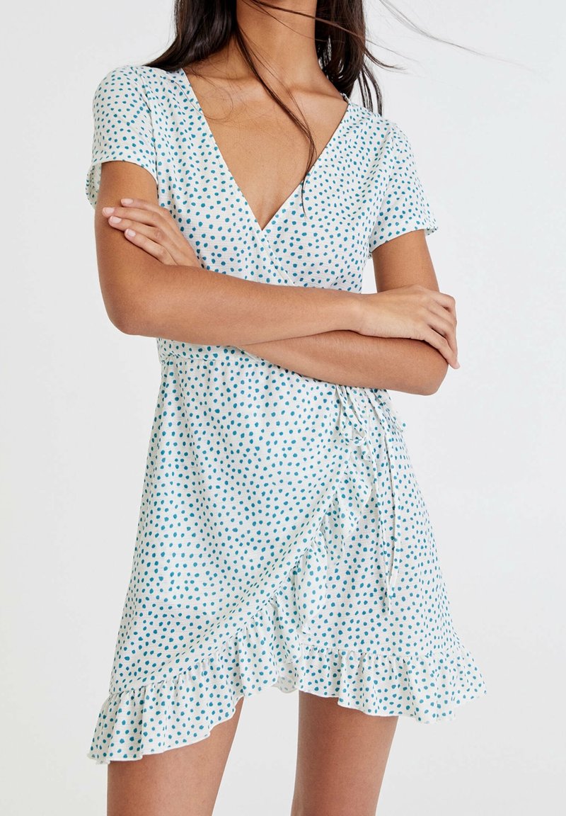 Femme portant une robe portefeuille blanche à pois bleus avec un ourlet volanté, debout les bras croisés devant un fond uni.