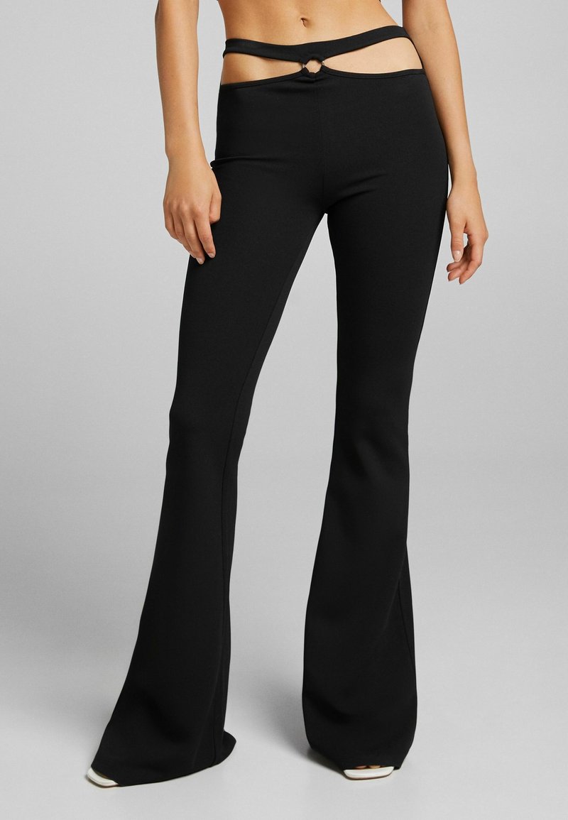 Personne portant un pantalon noir évasé taille haute avec une taille découpée ornée d'un anneau argenté, debout devant un fond gris uni.
