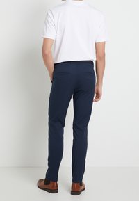 Marina smal passform byxor med en slät textur, bälteshälla och två bakfickor. Kombinerat med en vit T-shirt och bruna skor.