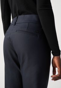 Tailored navy trousers with a smooth texture, featuring a back pocket and an embossed logo. Worn with a black long-sleeve top.
