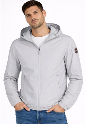 Mand iført lysegrå hættejakke med lynlås og elastiske manchetter, hænder i lommerne, hvid t-shirt og blå jeans på hvid baggrund.