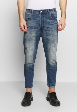 Mężczyzna ubrany w wyblakłe, znoszone, obcisłe niebieskie jeansy z podwiniętymi nogawkami oraz czarne sznurowane buty, stojący na jednolitym tle.