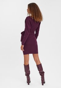 Robe tricotée en violet profond avec des manches longues, une taille ceinturée et une silhouette ajustée, associée à des bottes bouffantes jusqu'aux genoux dans une couleur similaire.